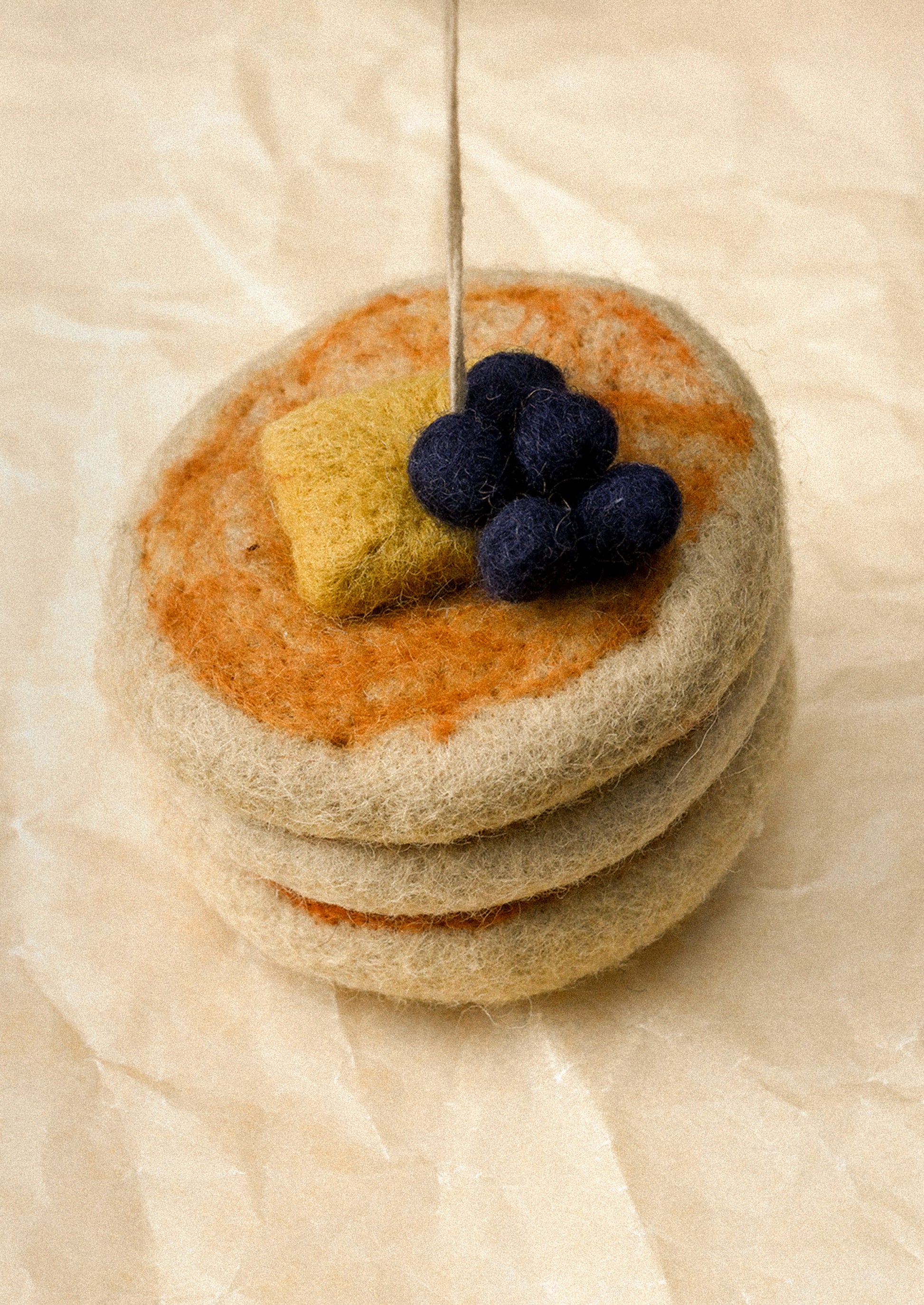 A felted wool miniature stack of three pancakes topped with orange-brown syrup, a yellow butter piece, and dark blue felted blueberries, with a metal pin sticking up from the center, set on a beige textured paper background.