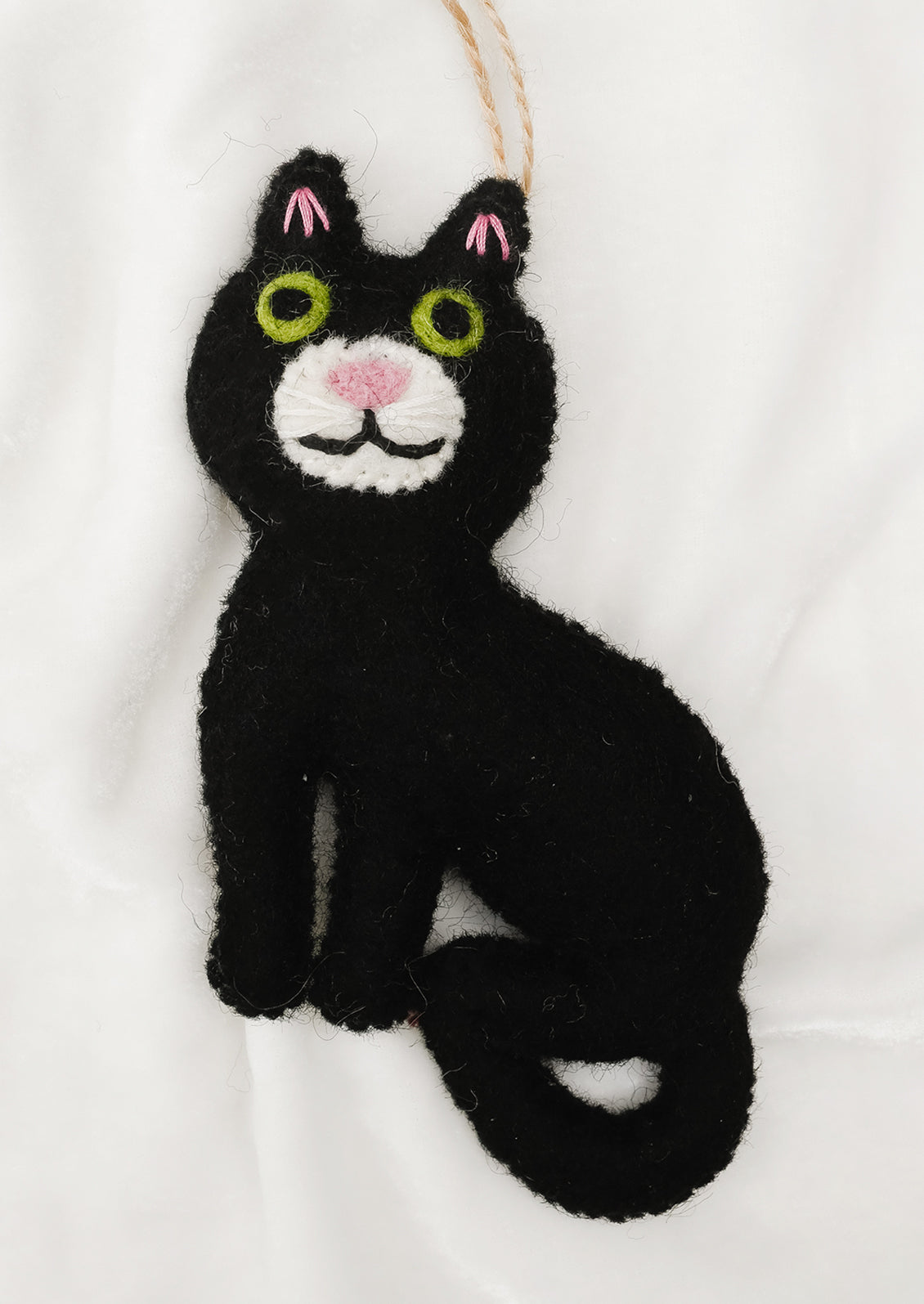 A black felt cat ornament with embroidered green eyes, pink nose and ear accents, and a white chin, hanging from a tan string against a white fabric background.