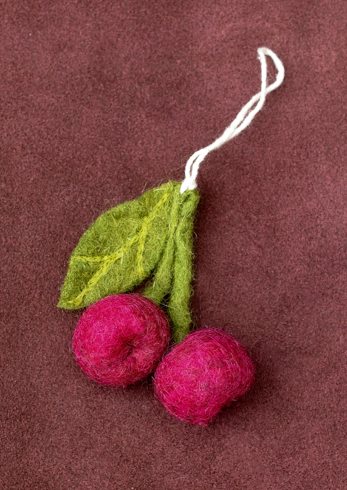 A felted wool ornament shaped like two pinkish-red cherries with green leaves and a white hanging string, photographed on a dark brown textured background.