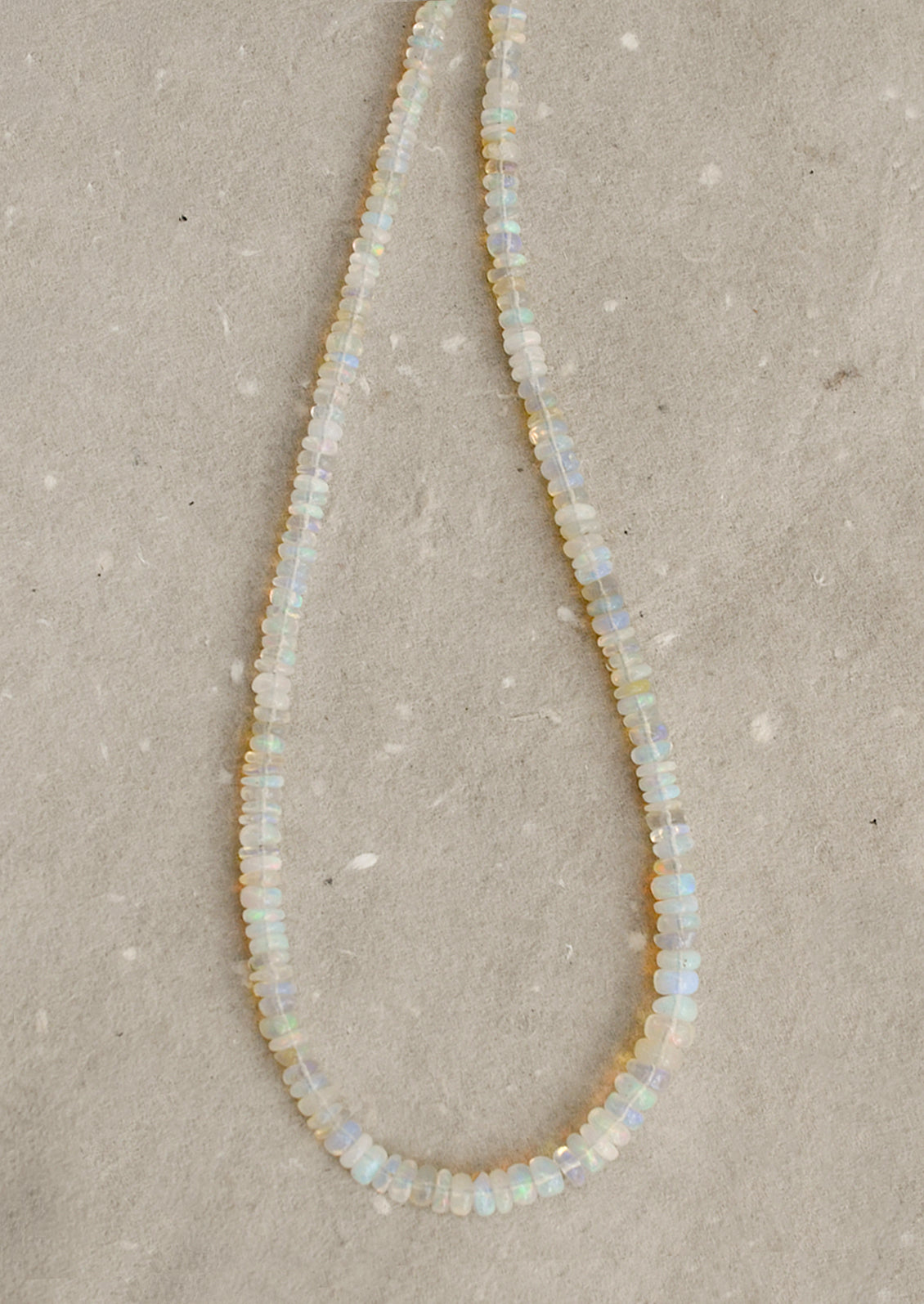 A beaded necklace made of small, irregular opal-like beads that shimmer with iridescent colors of white, pale blue, and soft yellow, laid out on a textured beige surface.