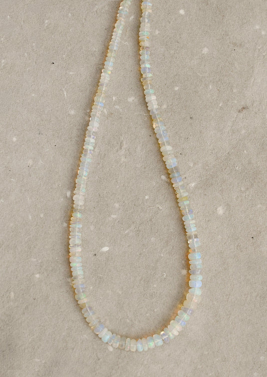 A beaded necklace made of small, irregular opal-like beads that shimmer with iridescent colors of white, pale blue, and soft yellow, laid out on a textured beige surface.