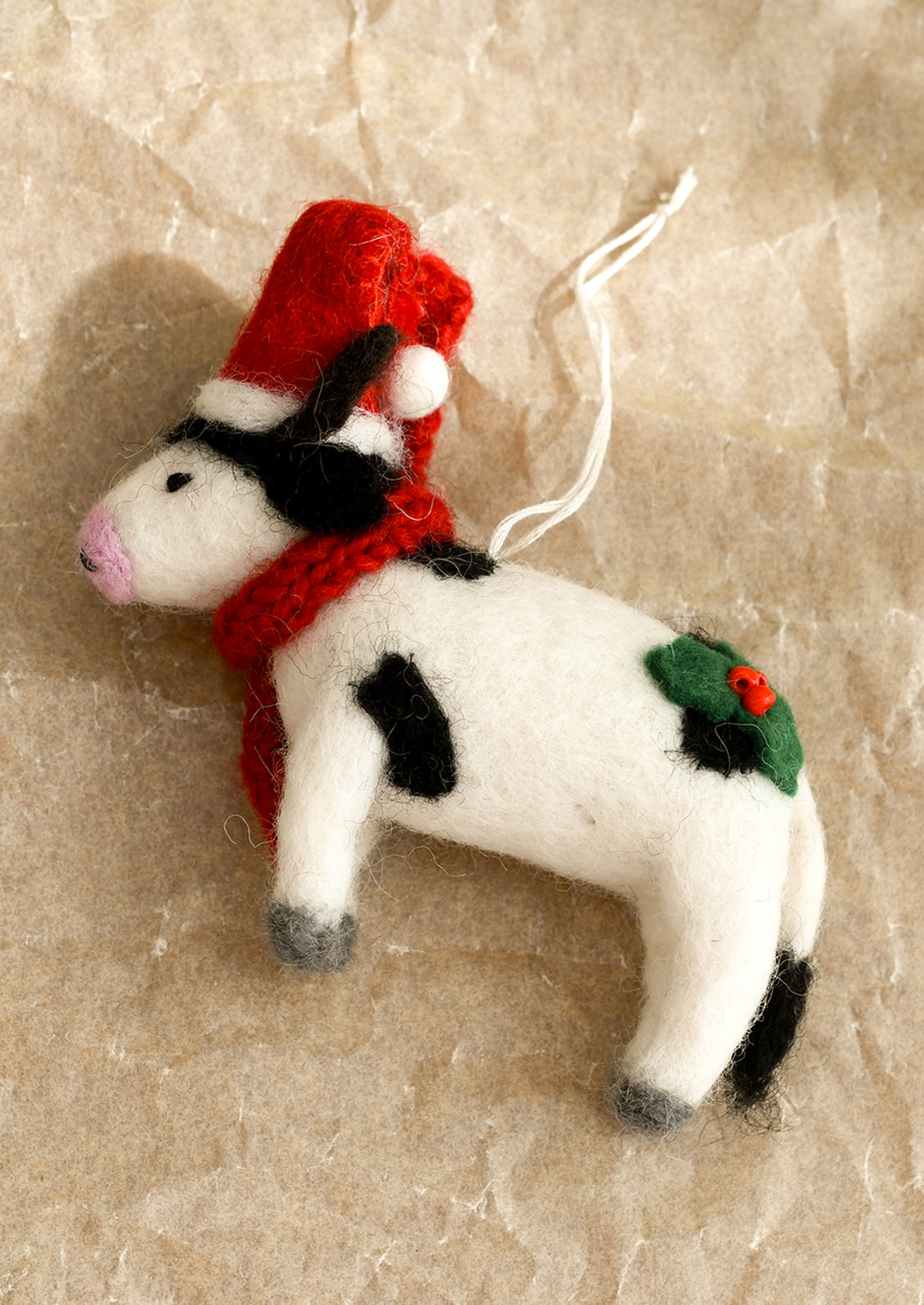 A felted wool ornament in the depiction of a cow, wearing red scarf and santa hat.