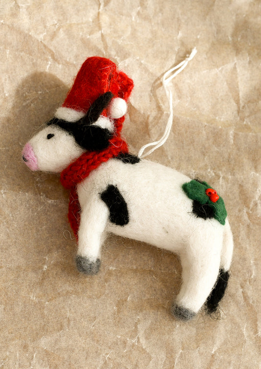 A felted wool ornament in the depiction of a cow, wearing red scarf and santa hat.