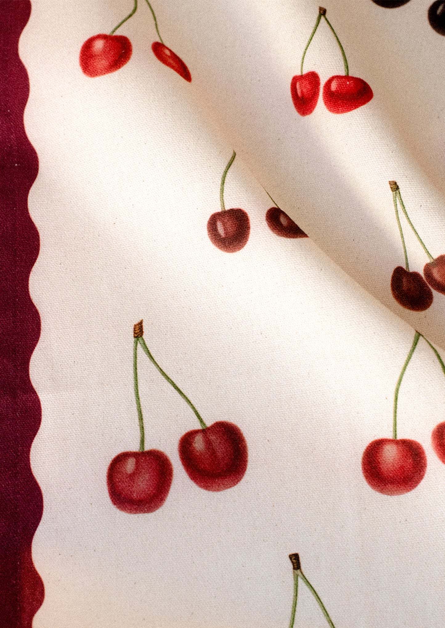 Close-up of a white cotton tea towel with red cherry illustrations and green stems, with a red scalloped border at one edge.