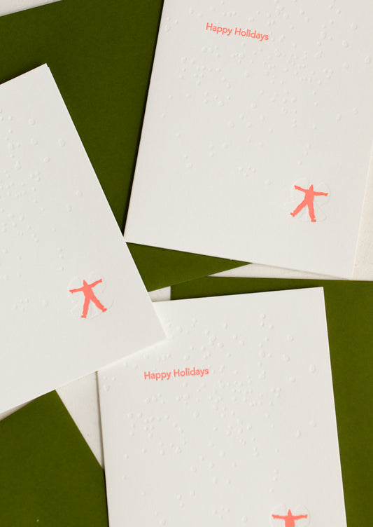 White letterpress greeting cards with neon 'Happy Holidays' text and red silhouette of a person making snow angels.