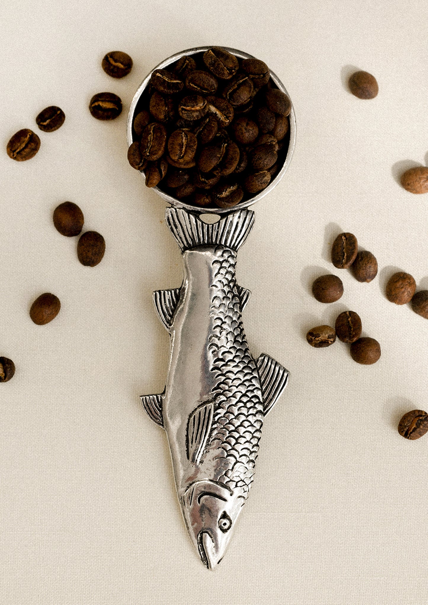 Silver fish-shaped coffee scoop with coffee beans on a light background