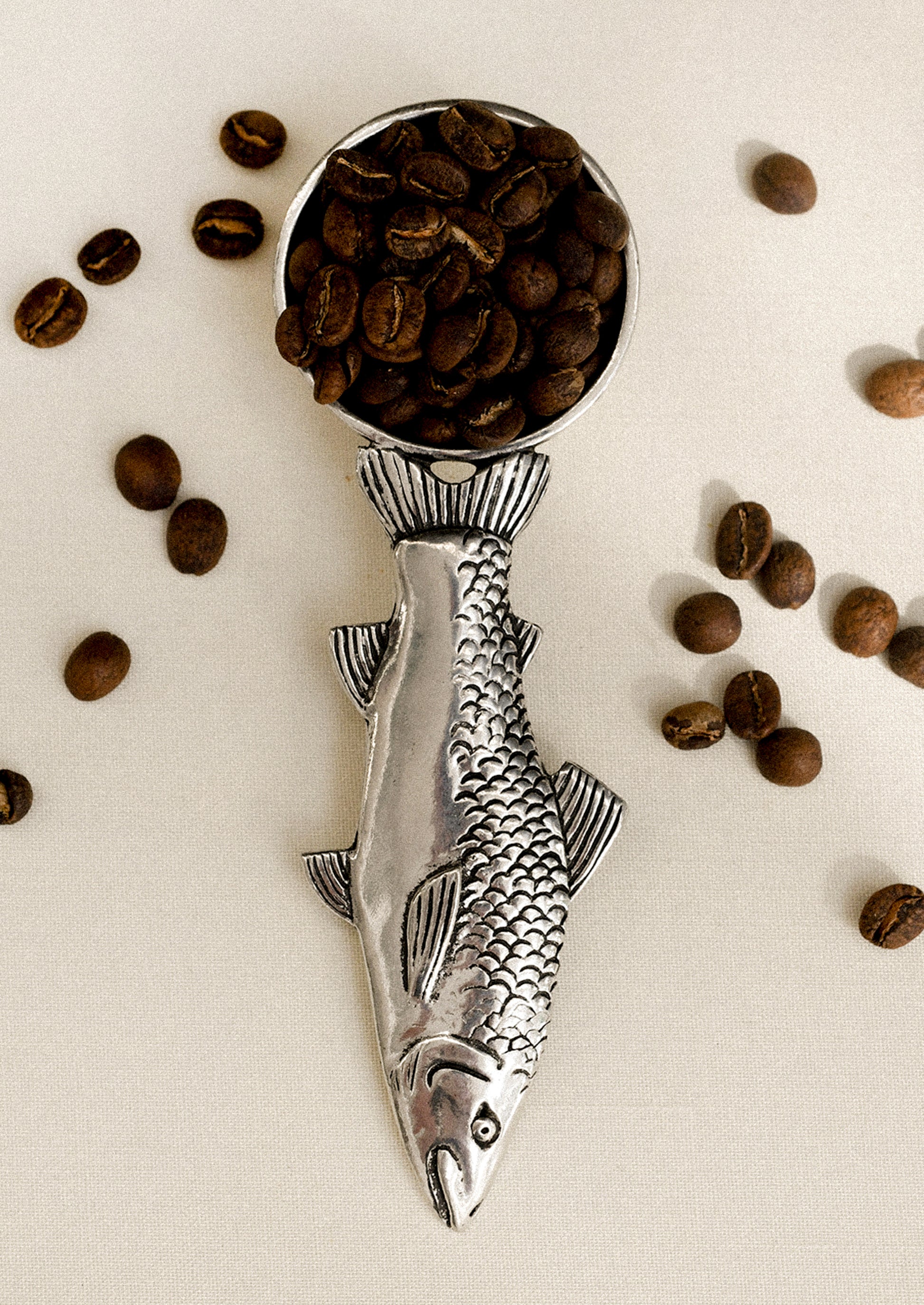Silver fish-shaped coffee scoop with coffee beans on a light background