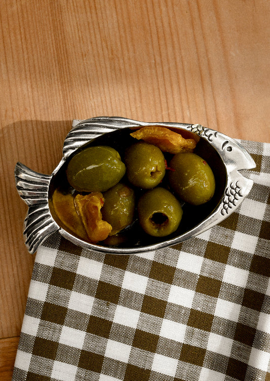 Metallic fish-shaped bowl with green olives and bread on a checkered cloth.