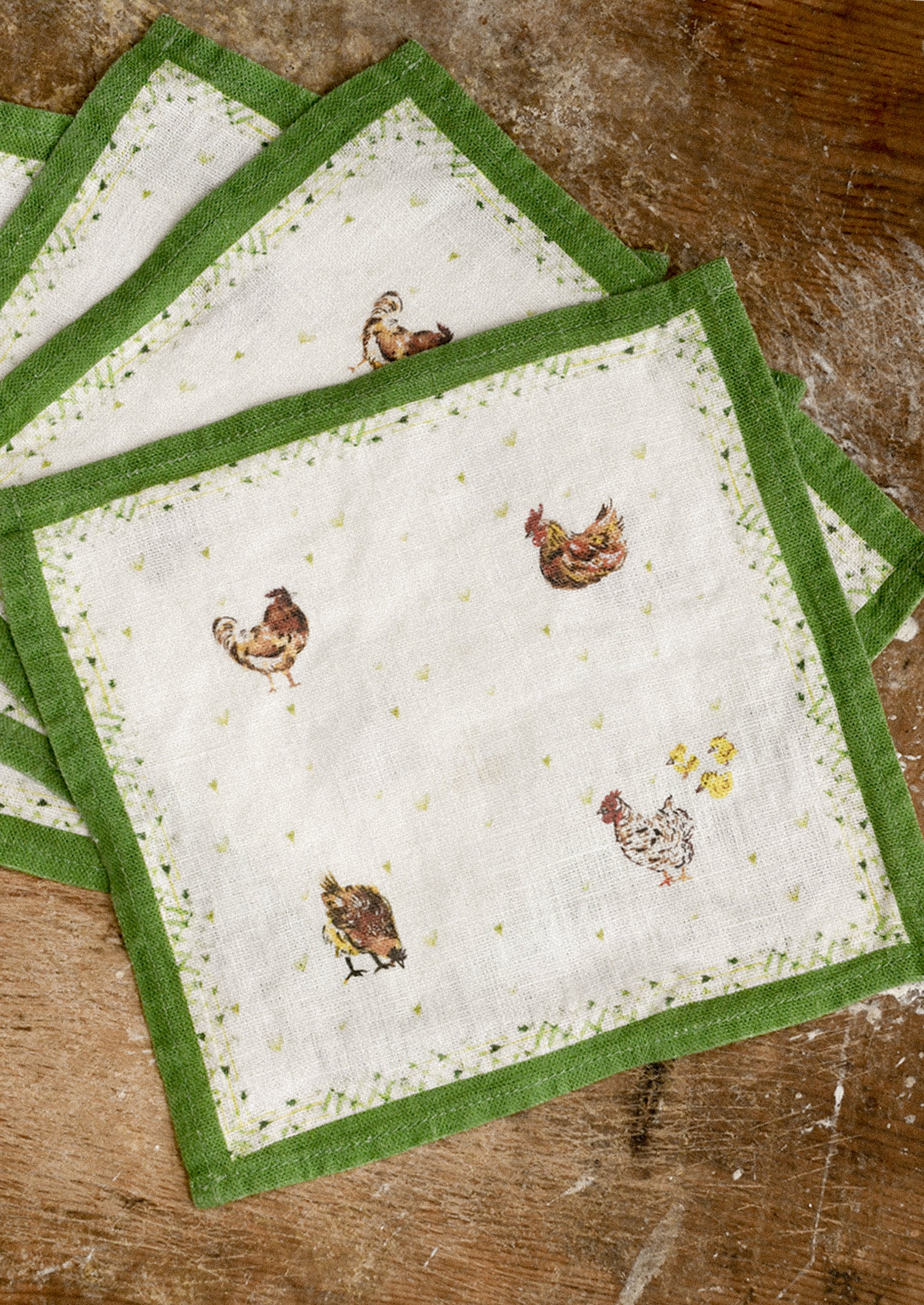 Set of four cocktail napkins with green borders and chicken designs on a wooden surface
