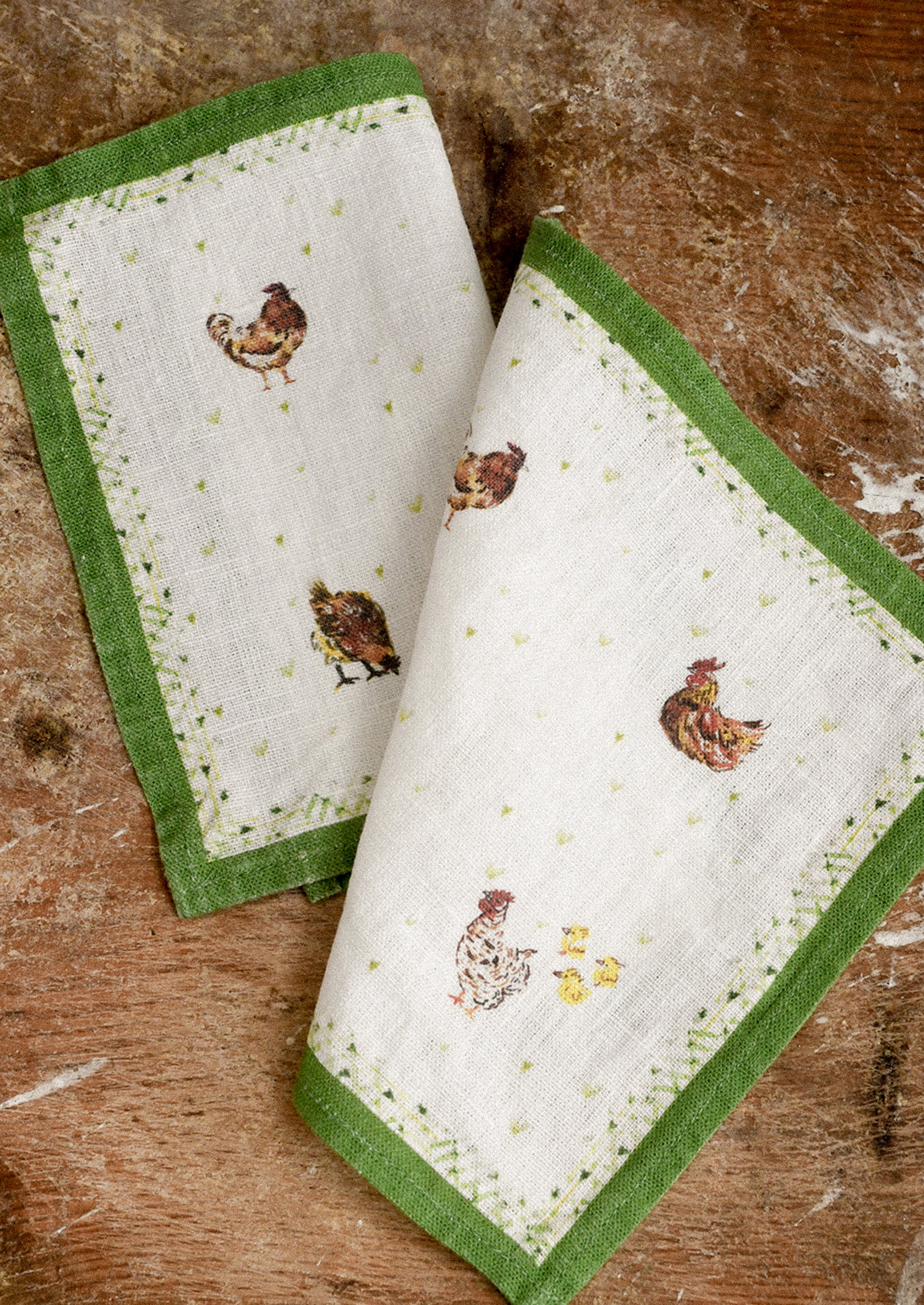 Set of four cocktail napkins with green borders and chicken designs on a wooden surface