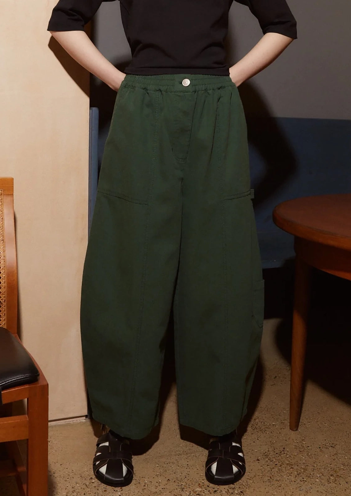 A person wearing dark green wide-leg cargo pants with a black top and black sandals, standing indoors with hands on hips.