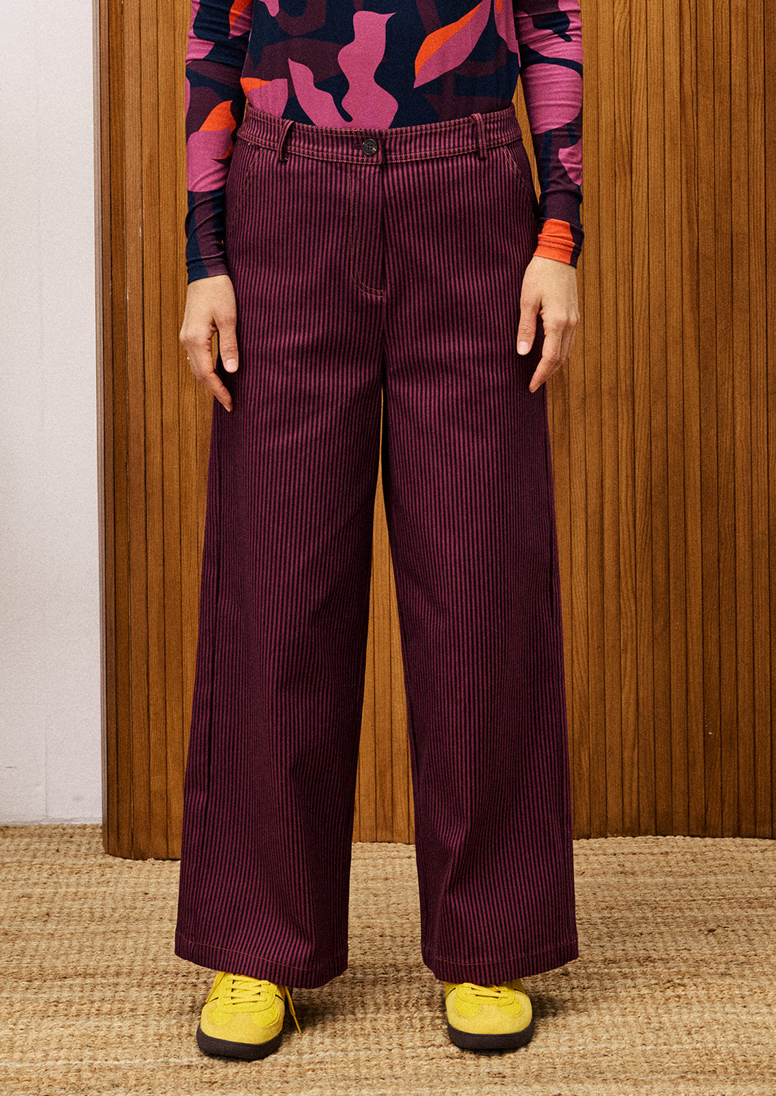Person wearing maroon corduroy pants with a colorful top and yellow shoes against a wooden panel background.