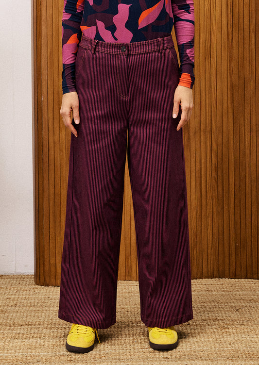 Person wearing maroon corduroy pants with a colorful top and yellow shoes against a wooden panel background.