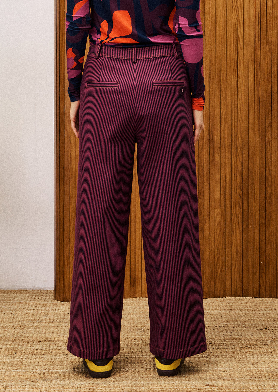 Person wearing a colorful top and purple striped pants standing against a wooden wall.