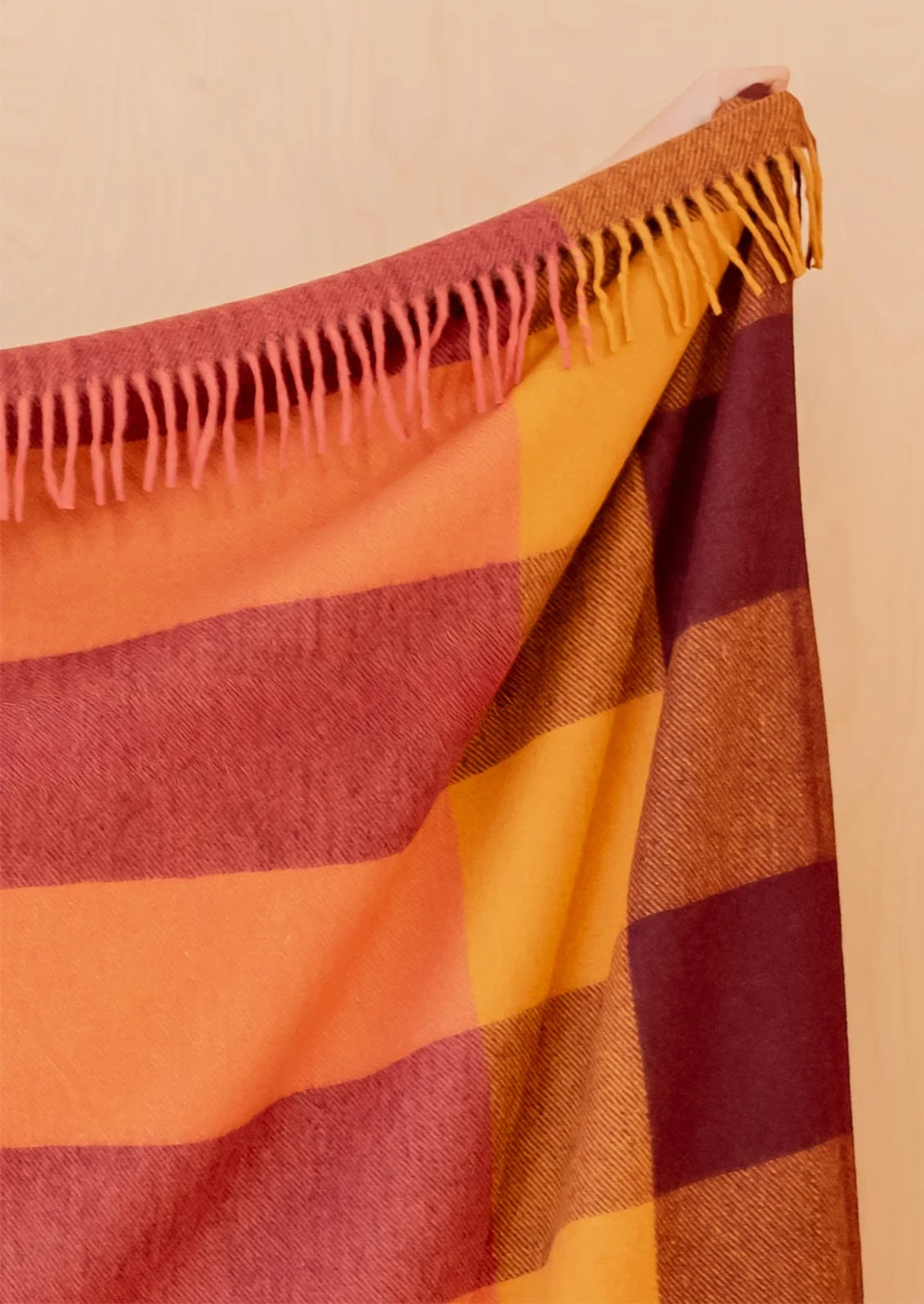 A close-up of a woven throw blanket with a warm plaid pattern in orange, rust red, mustard yellow, and brown, featuring fringed edges.