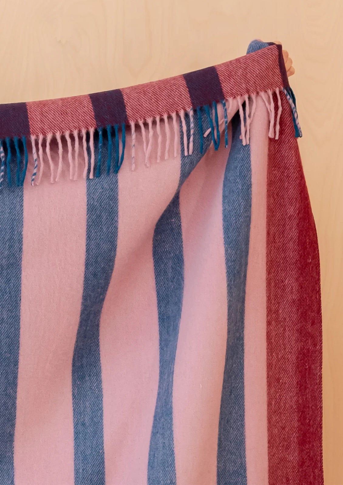 A close-up of a striped woven throw with fringe, shown hanging against a light wood background. The color pattern features vertical stripes in dusty pink, slate blue, and deep red, with dark purple stripes at the top edge.