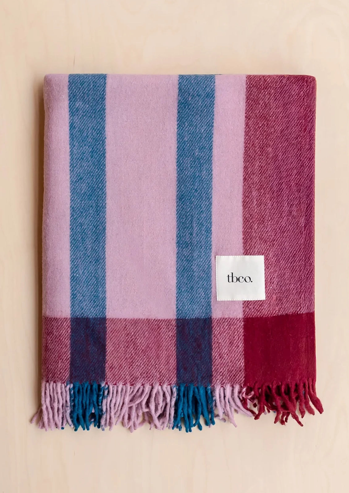A folded plaid throw blanket with vertical stripes in dusty pink, blue, and deep magenta, finished with fringed edges and a small white 'theco.' label, displayed on a light wood background.
