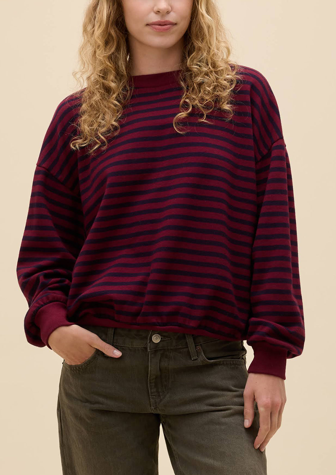 A woman wearing a dark burgundy and black horizontally striped crewneck sweatshirt with dark olive green jeans, shown from the waist up against a solid beige background.