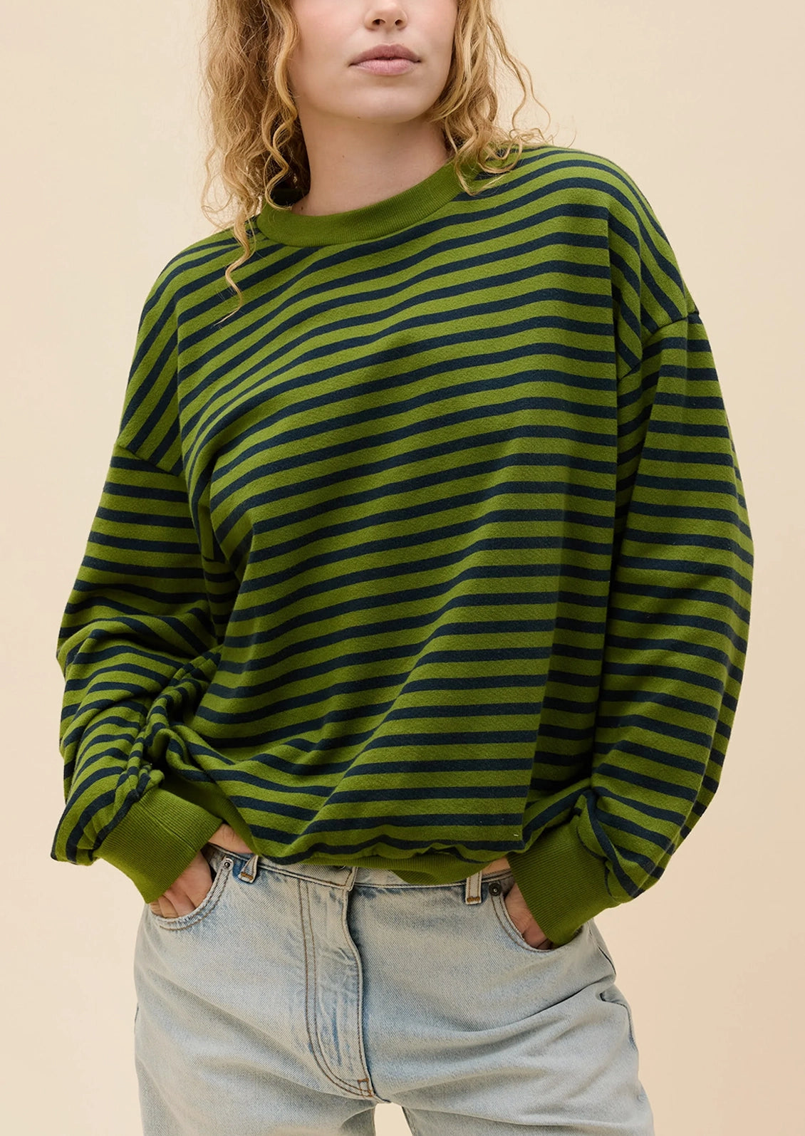A woman wearing an oversized long-sleeve crewneck sweater with horizontal olive green and dark green stripes, paired with light-wash jeans, against a solid beige background.