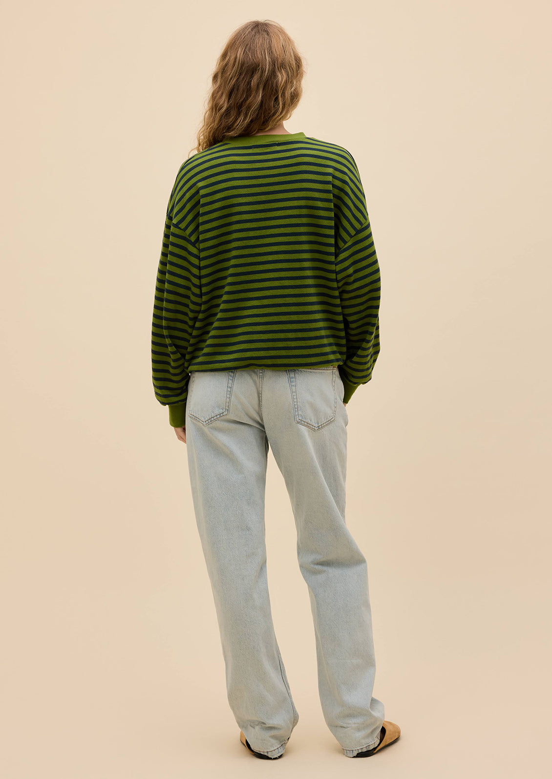 A person shown from the back wearing a green and dark green horizontally striped long-sleeve knit sweater, light-wash wide-leg jeans, and tan shoes, against a solid beige studio background.