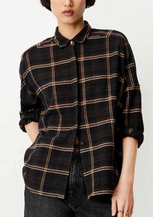 A woman wearing a black plaid button-down shirt with rolled-up sleeves and dark jeans, photographed against a plain light background.