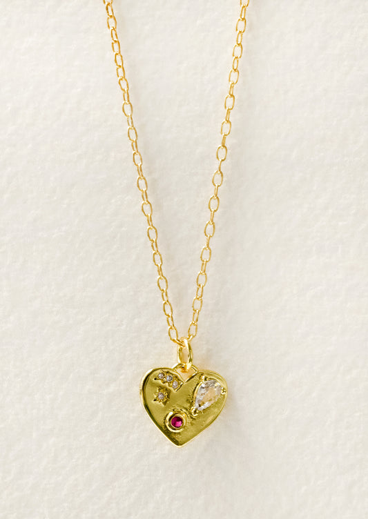 A gold necklace with a gold heart charm adorned with glass crystal and a red gemstone.
