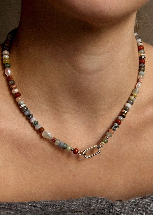 A woman wearing a gold carabiner necklace adorned with various colored gemstones in muted tones.