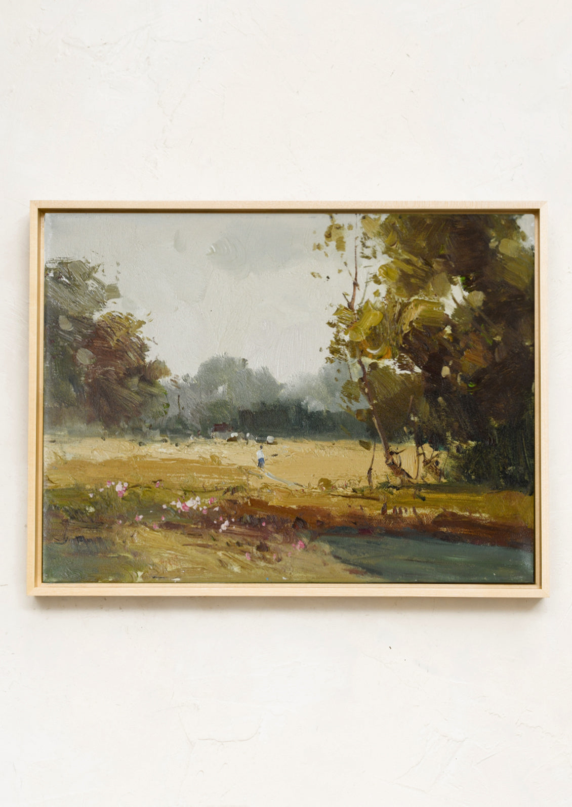 A framed landscape oil painting on canvas, featuring a grassy field, trees, and a stream, housed in a light natural wood frame and mounted on a textured off-white wall.