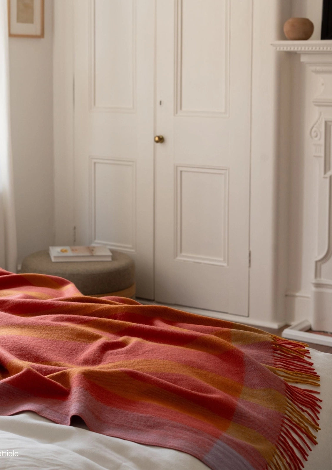 A cozy bedroom scene featuring a soft striped throw blanket draped over a bed. The blanket has a warm color palette of terracotta red, orange, and yellow stripes with fringed edges.