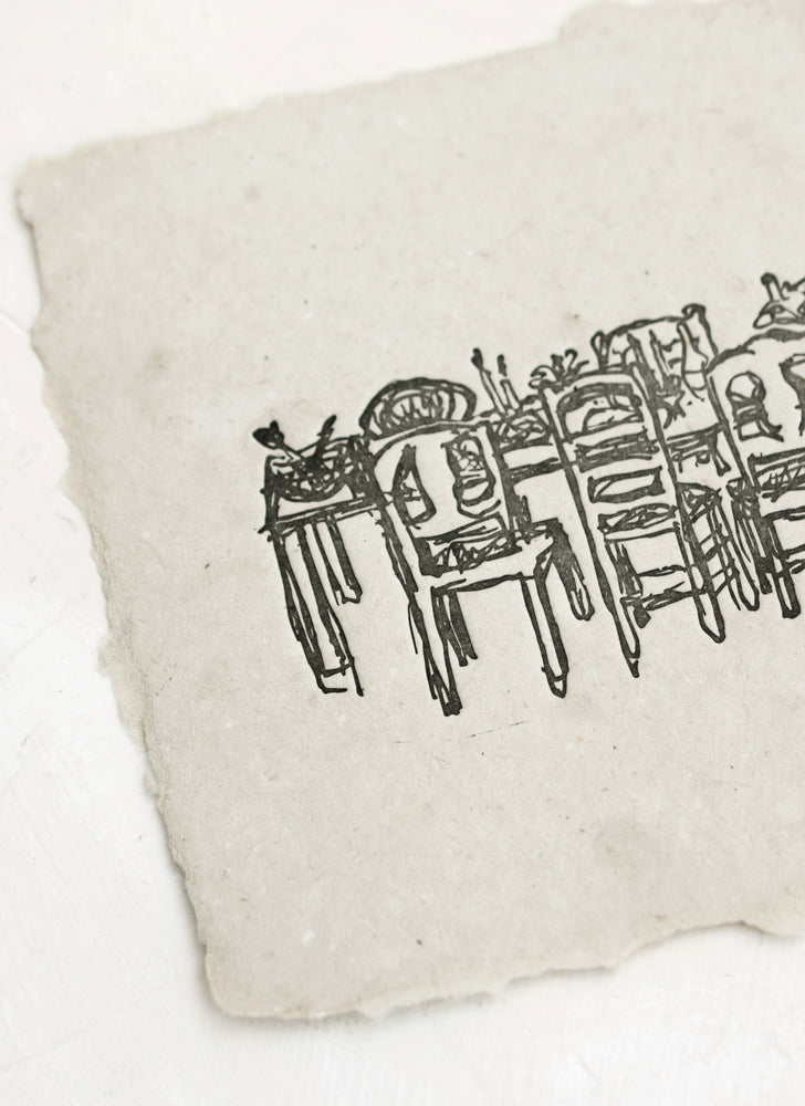 An art print made from grey handmade paper with letterpressed image of a dining table with chairs.