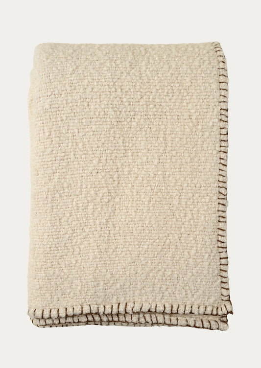 A folded beige textured throw blanket with a whipstitched border in brown.