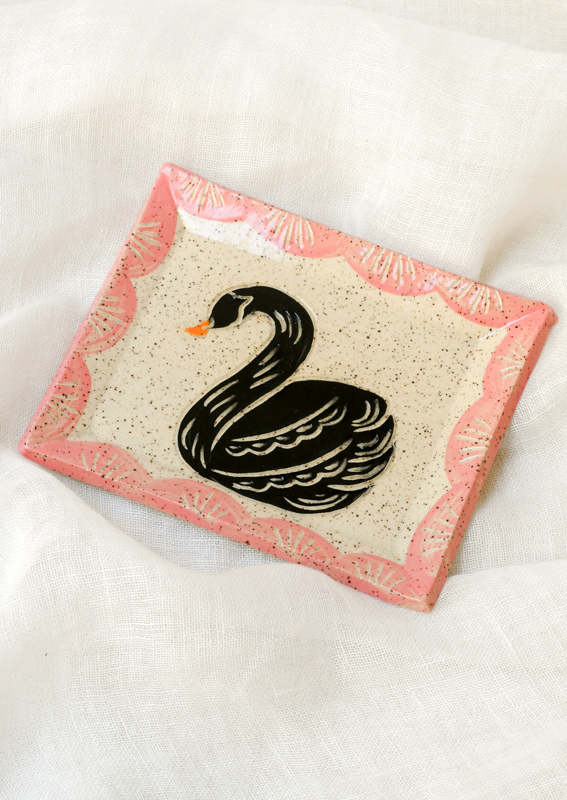 A small rectangular ceramic catchall tray with an etched black swan in the center and a pink painted scallop border along the edges.