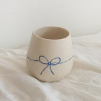 2: A white rounded ceramic cup with hand painted blue bow.