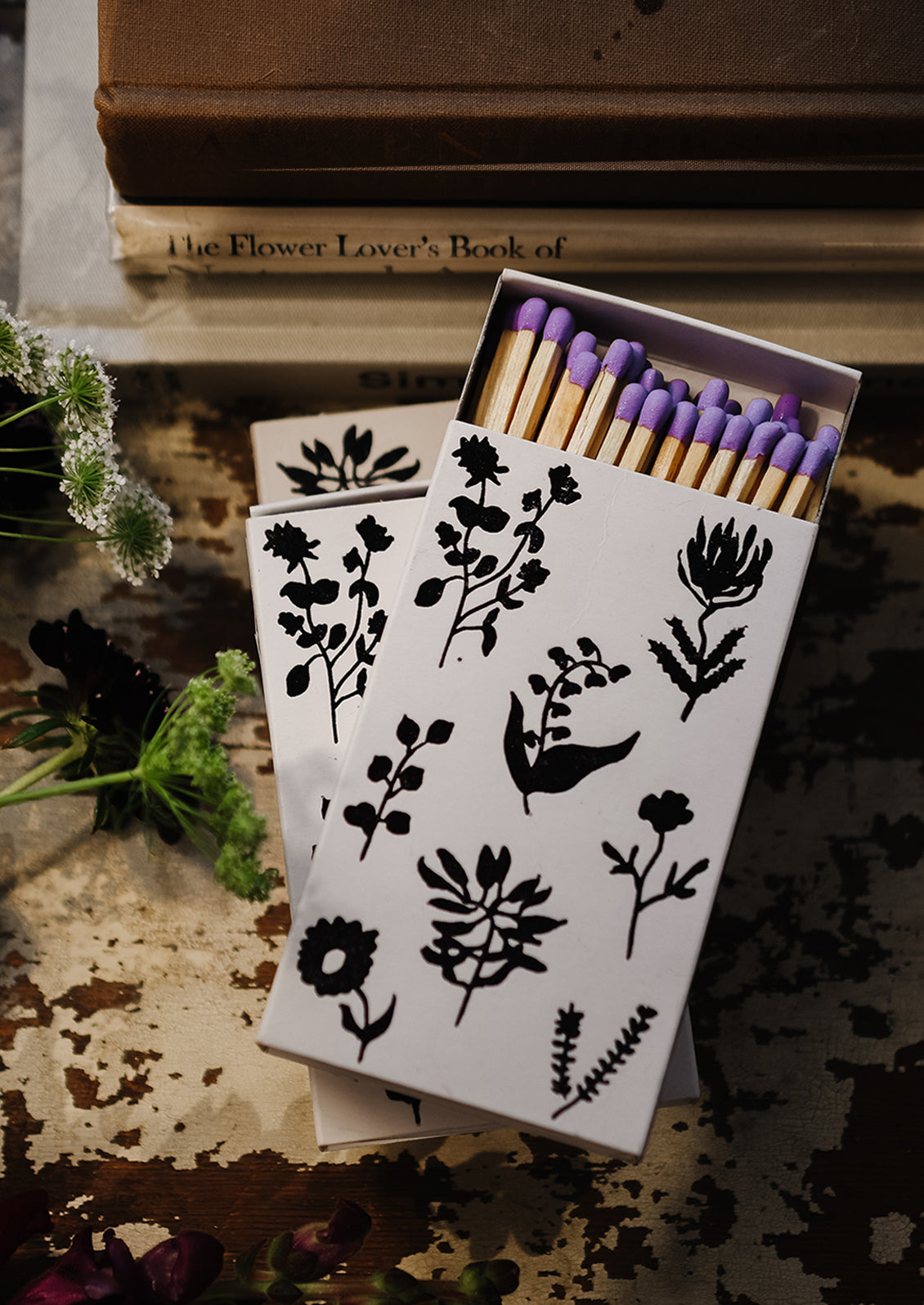 A matchbox in white with black botanical print, housing purple tipped matches.