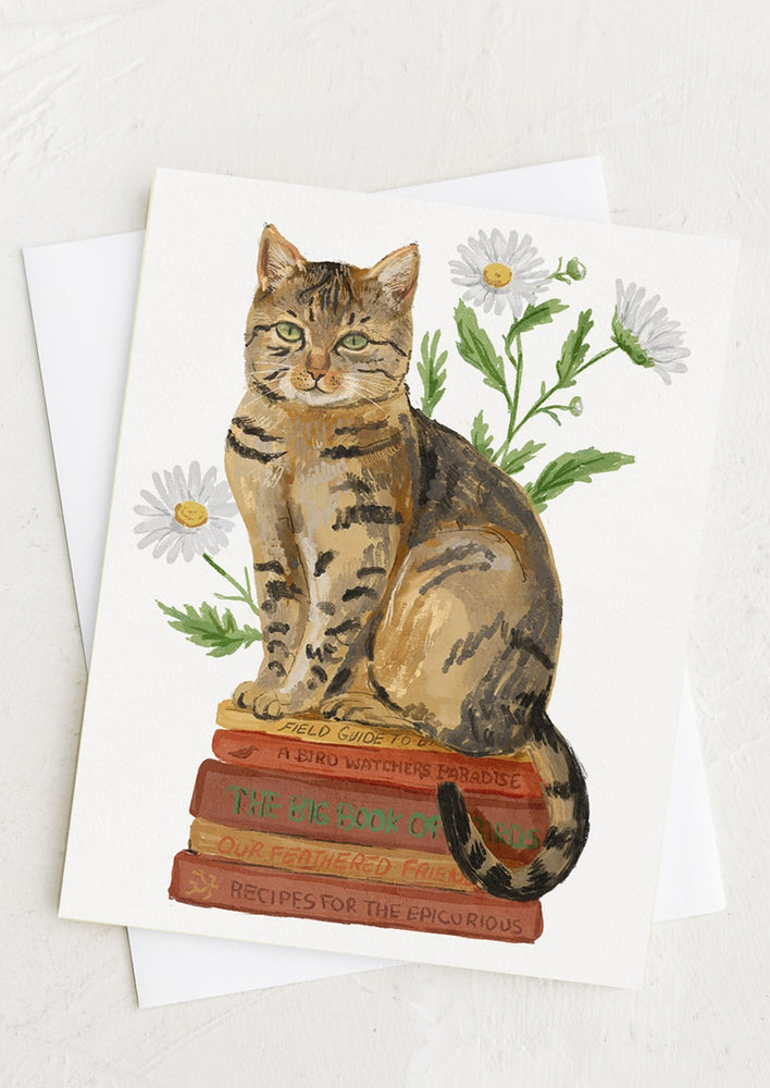 A greeting card with illustration of cat sitting on pile of books.