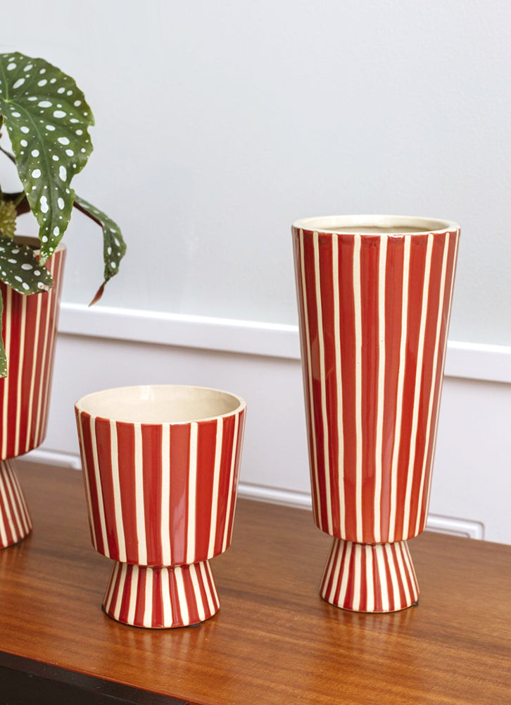 A tall and skinny vase with conical shape in red ceramic with vertical thin white stripes.