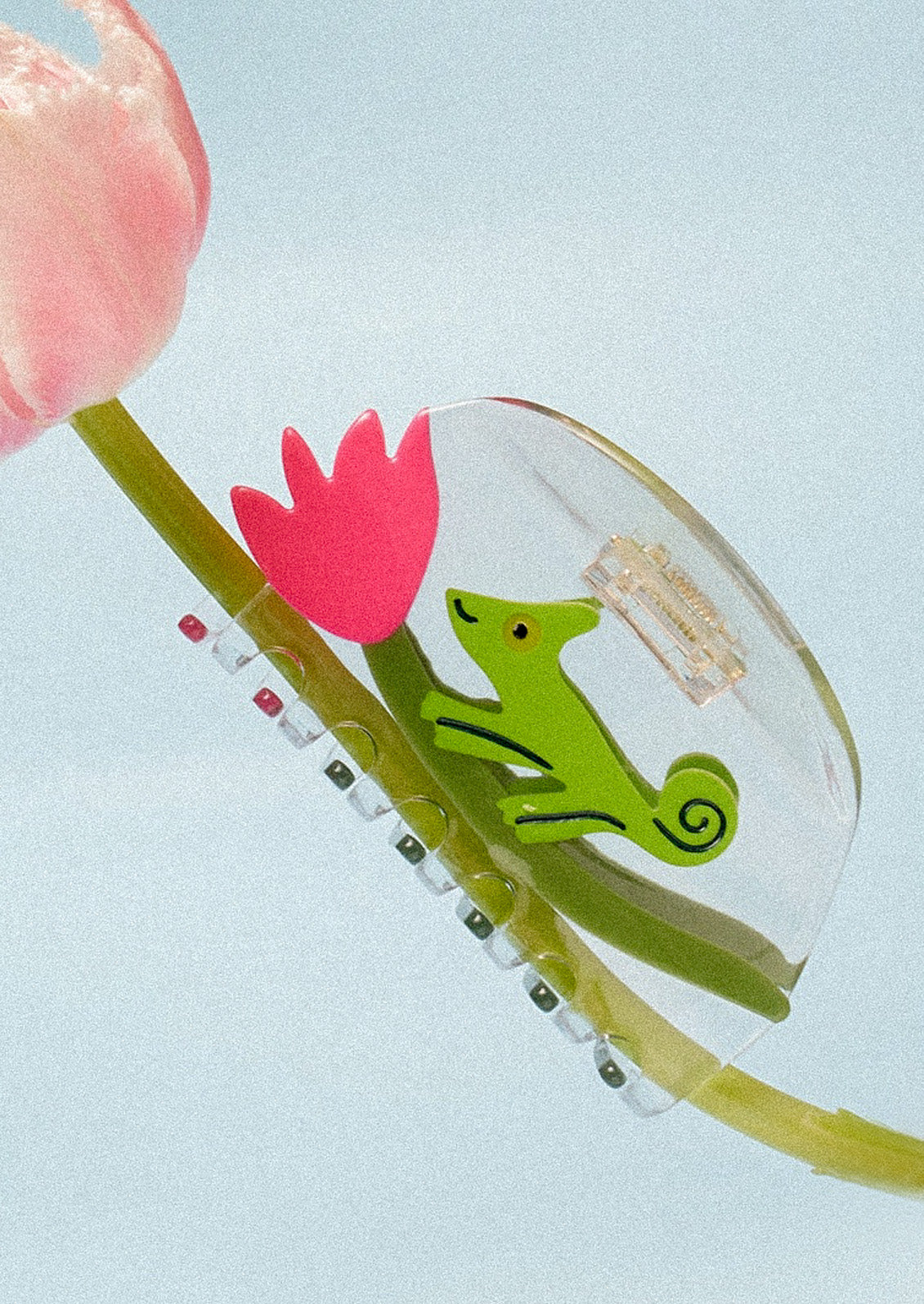 A clear claw clip with a green chameleon and pink tulip attached to the stem of a pink flower on a blue background.