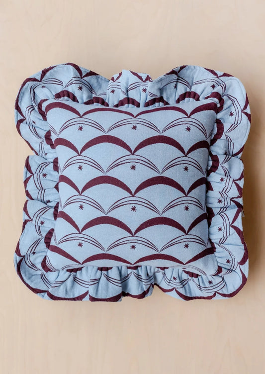 A square decorative throw pillow with a ruffled border, featuring a light blue and burgundy scallop pattern on a light wood background.