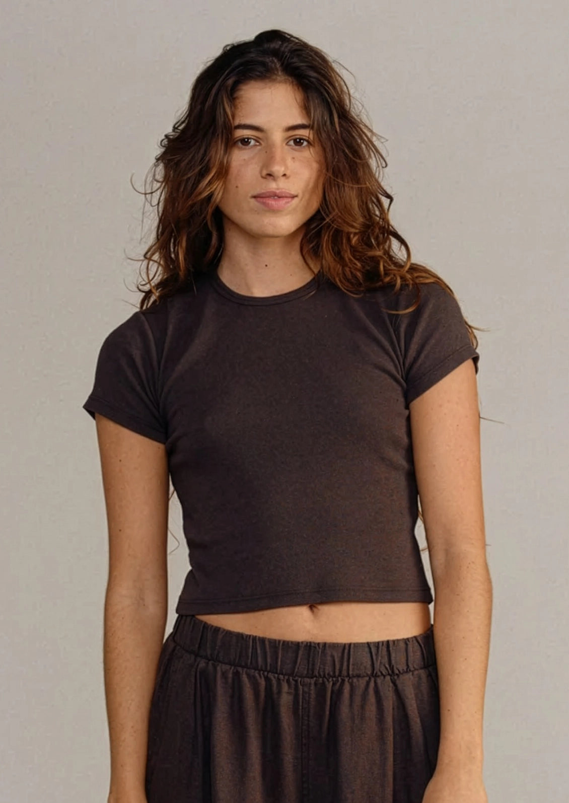 A woman modeling a dark brown cropped short-sleeve t-shirt and matching dark brown pants against a neutral gray studio background.