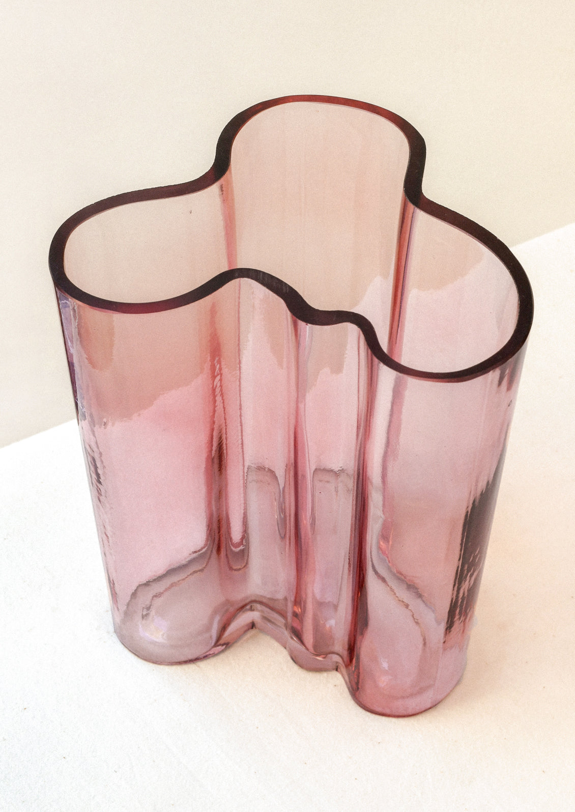 A glass vase with a wavy, curvy overall shape, in transparent rose hue.