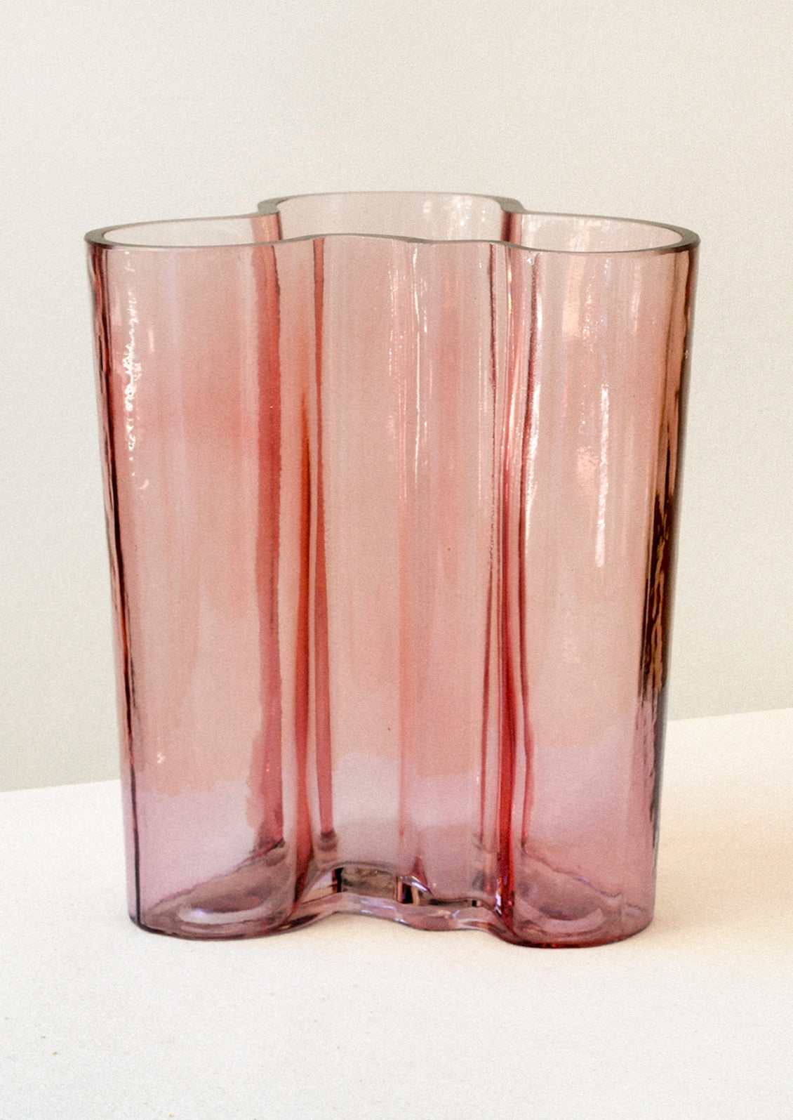 A glass vase with a wavy, curvy overall shape, in transparent rose hue.