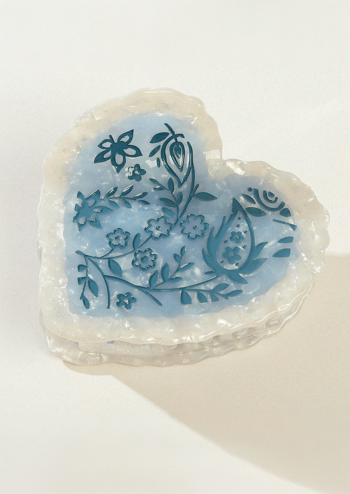 A large heart shaped hair claw with a pearlescent border and a blue floral design in the middle.