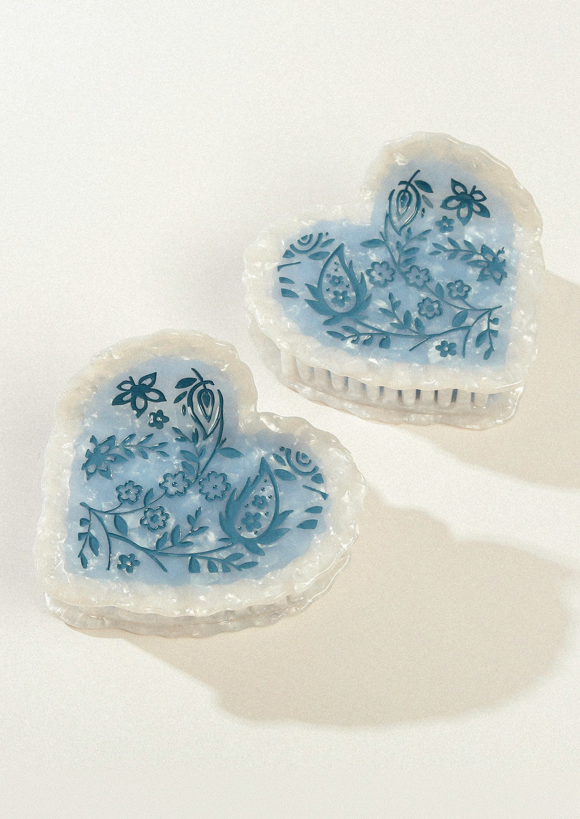 Two large heart shaped hair claws with a pearlescent border and a blue floral design in the middle.