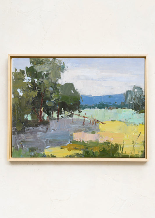 A framed landscape oil painting featuring green trees, a pale blue sky, and a bright yellow field, hung on a textured off-white wall.