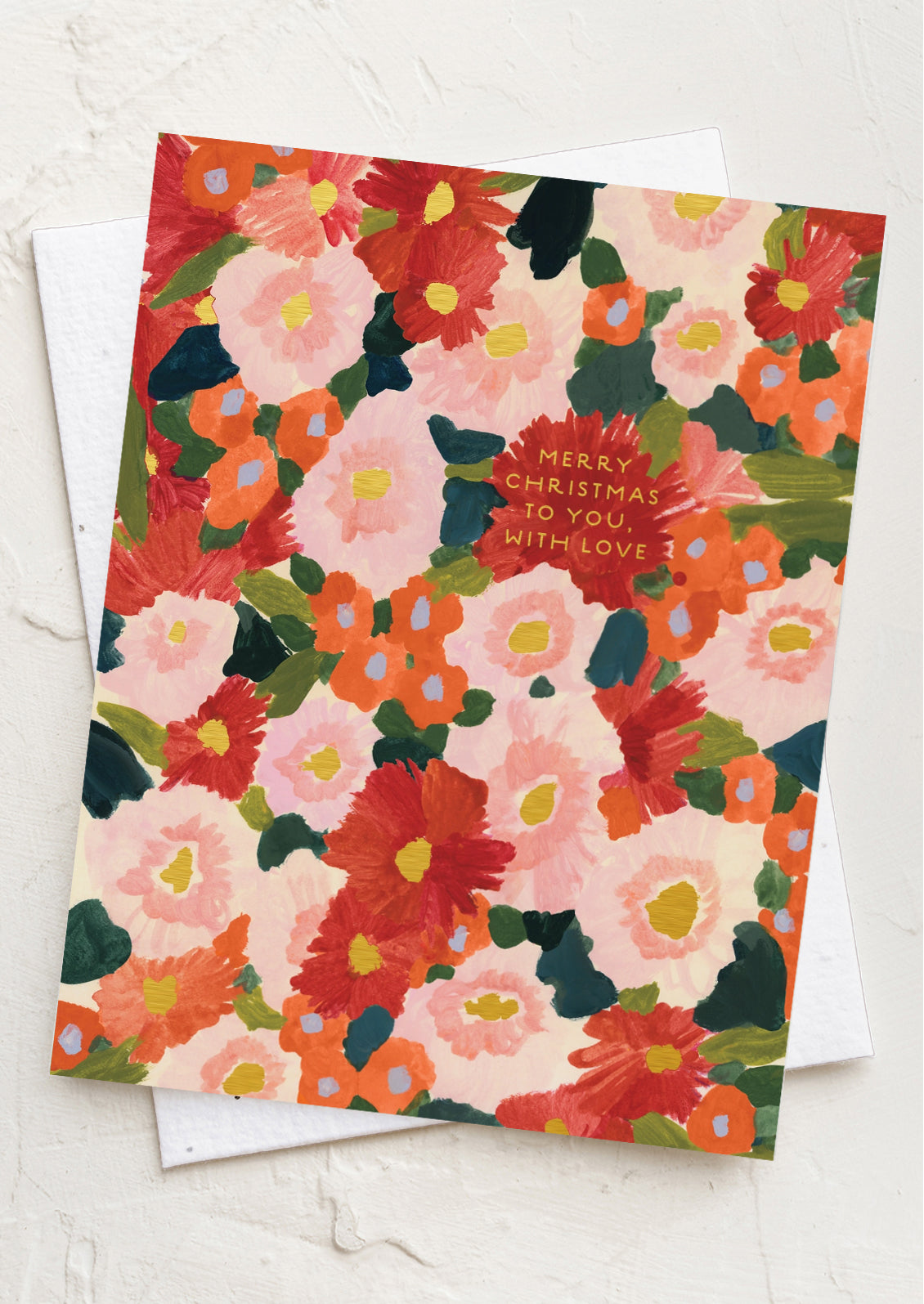 A floral Christmas greeting card with a warm multicolor pattern of red, orange, pink, and green flowers on a cream background, layered over a white envelope on a textured white surface.