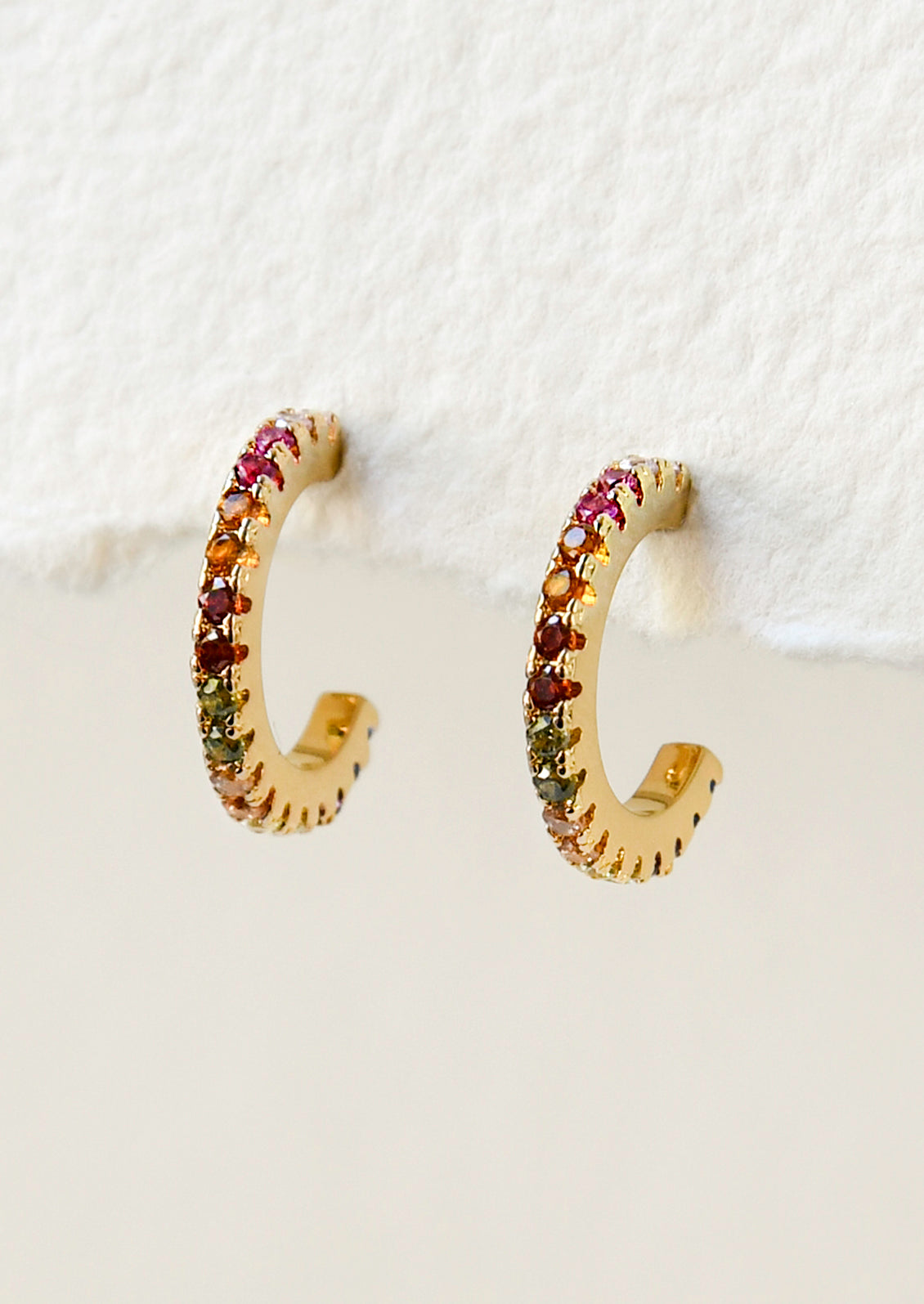 A pair of gold hoop earrings with pink, range, red, green, and clear gems in a gradient design.