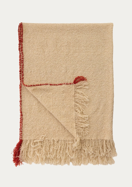 A folded beige woven throw blanket with fringed edges and a contrasting red tassel detail, shown on a light gray background.