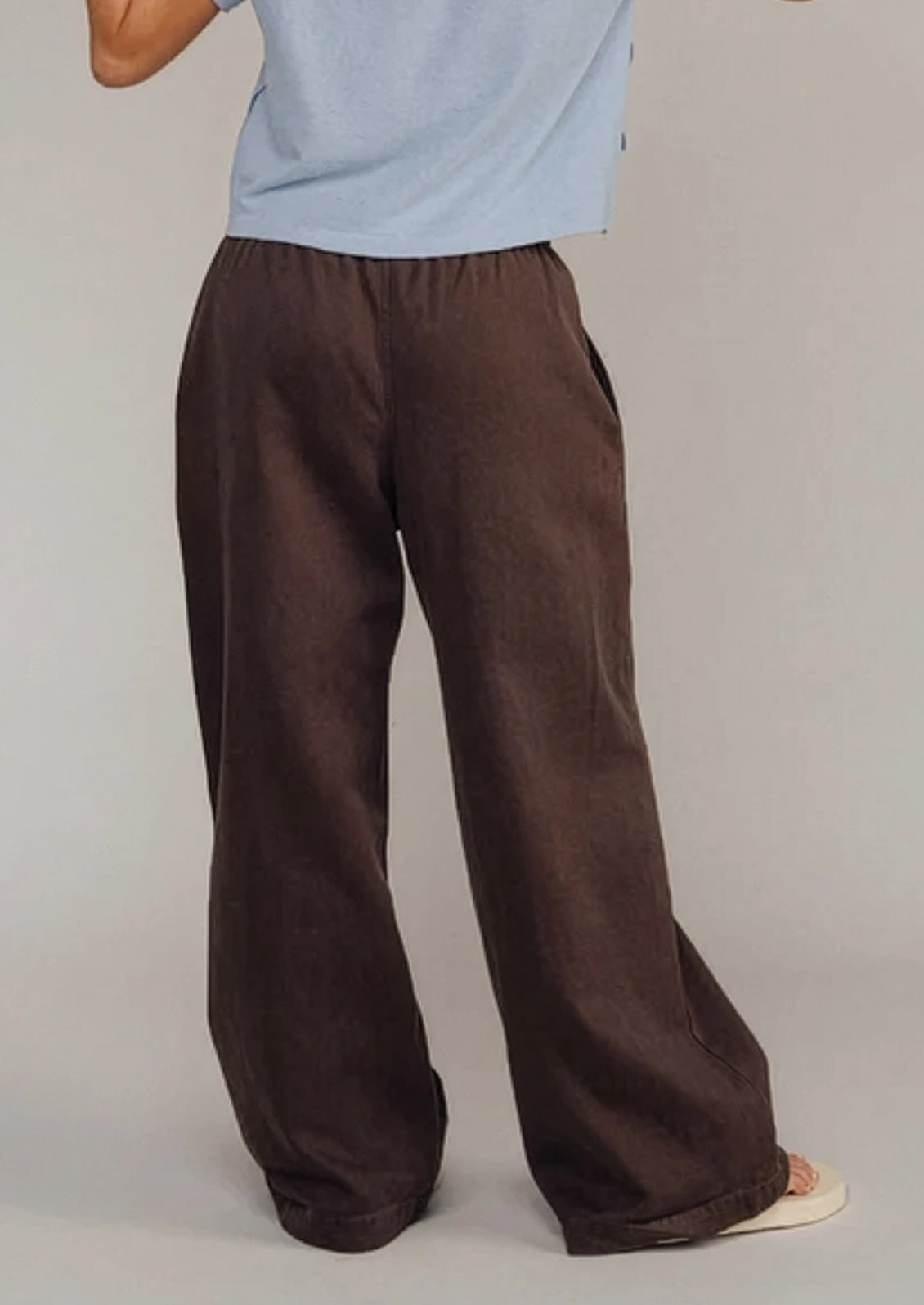 A person wearing loose, wide-leg brown pants with a light blue top, shown from the back against a plain light gray studio background.