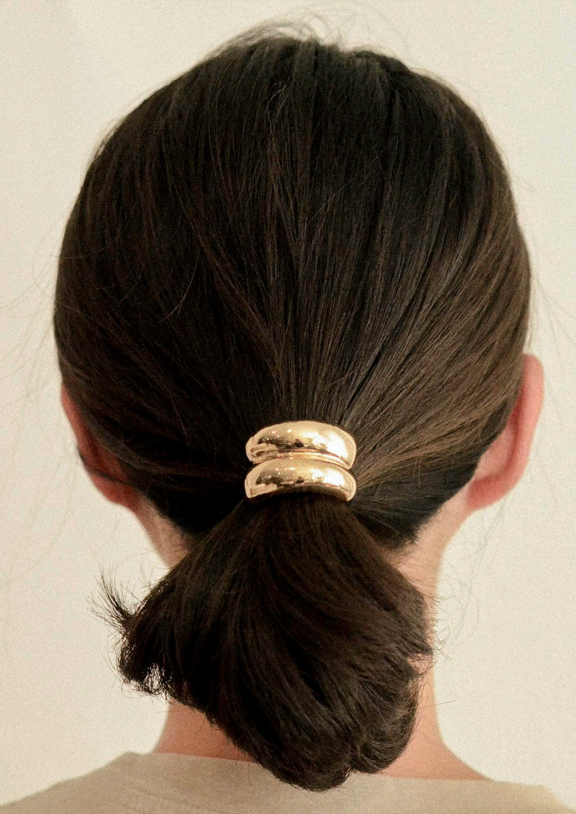 A woman wearing a black elastic hair tie with a gold metal pendant with a double arch design holding her hair in a ponytail.