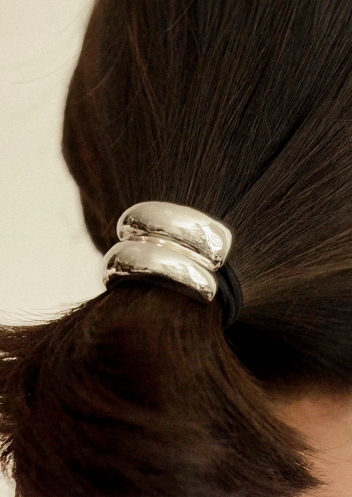 A woman wearing a black elastic hair tie with a silver metal pendant with a double arch design holding her hair in a ponytail.